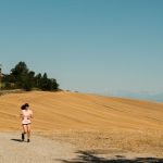 A woman runs on a gravel path through a sunlit rural landscape with dry fields and blue sky.